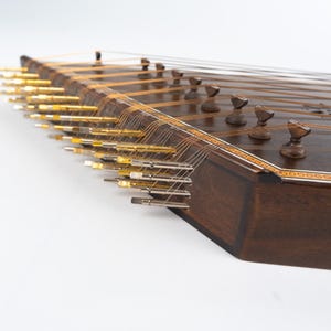 May include: A dark brown wooden santur, a hammered dulcimer, with gold tuning pegs and strings. The instrument has a decorative inlaid border.