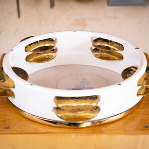 May include: White  doumbek with gold cymbals. The doumbek has a white finish and a gold rim. The cymbals are attached to the side of the doumbek and are made of gold metal.