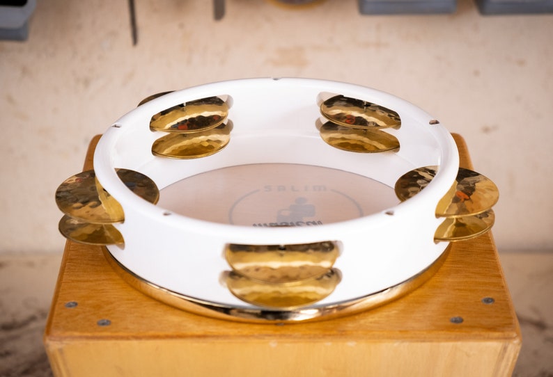 May include: White  doumbek with gold cymbals. The doumbek has a white finish and a gold rim. The cymbals are attached to the side of the doumbek and are made of gold metal.