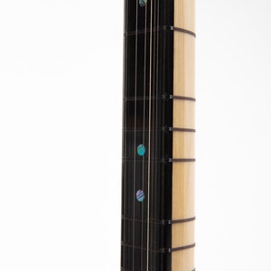 May include: Close-up of the fretboard of a musical instrument. The fretboard is made of wood and has a black finish. There are multiple strings running along the fretboard. The fretboard has several inlaid dots made of abalone shell.