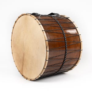 Op de afbeelding: Een grote, bruine houten trommel met een lichtbruin vel. De trommel heeft een donkerbruine houten body en is bespannen met zwart-wit touw. Er is een zwarte riem bevestigd om te dragen.