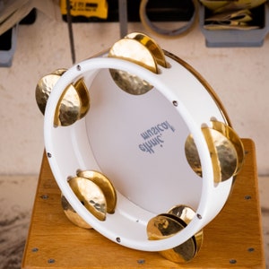 May include: White wooden frame with six gold metal cymbals, a percussion instrument called a  "Djembe". The frame has the text "Jhinic Musical" printed on it.