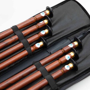 Puede incluir: Un conjunto de seis flautas de madera en una funda de transporte negra. Las flautas son marrones con veta de madera oscura y tienen detalles met&aacute;licos dorados. La funda tiene cierre de cremallera y forro de malla negra.