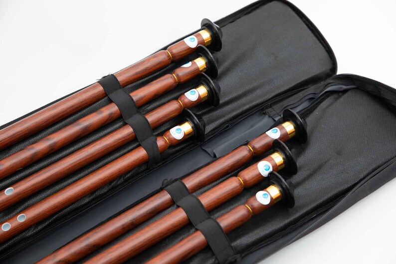 Puede incluir: Un conjunto de seis flautas de madera en una funda de transporte negra. Las flautas son marrones con veta de madera oscura y tienen detalles met&aacute;licos dorados. La funda tiene cierre de cremallera y forro de malla negra.