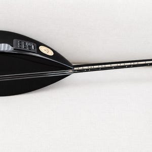 May include: A black saz, a long-necked stringed instrument, featuring a pear-shaped body with an integrated electronic control panel, a thin neck with metal frets, and multiple strings running along the length of the instrument to the headstock.