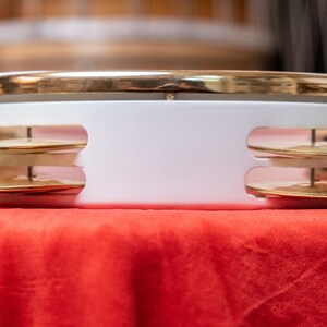 May include: A white tambourine with a gold rim and two sets of brass jingles. The tambourine is sitting on a red surface.