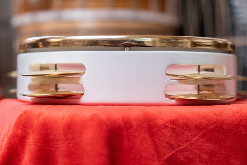 May include: A white tambourine with a gold rim and two sets of brass jingles. The tambourine is sitting on a red surface.