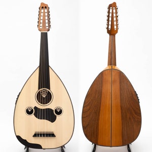 Könnte beinhalten: Eine hellfarbene, birnenförmige Oud mit dunklem Griffbrett und dekorativem Schallloch. Die Rückseite des Instruments besteht aus dunklen und hellen Holzpaneelen. Die Oud wird auf einem schwarzen Ständer vor weißem Hintergrund präsentiert.