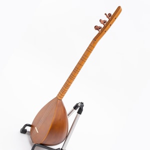May include: A wooden stringed instrument, likely a saz or similar folk instrument, with a pear-shaped body and a long neck. The instrument is a warm brown color and is resting on a silver and black stand against a white background.