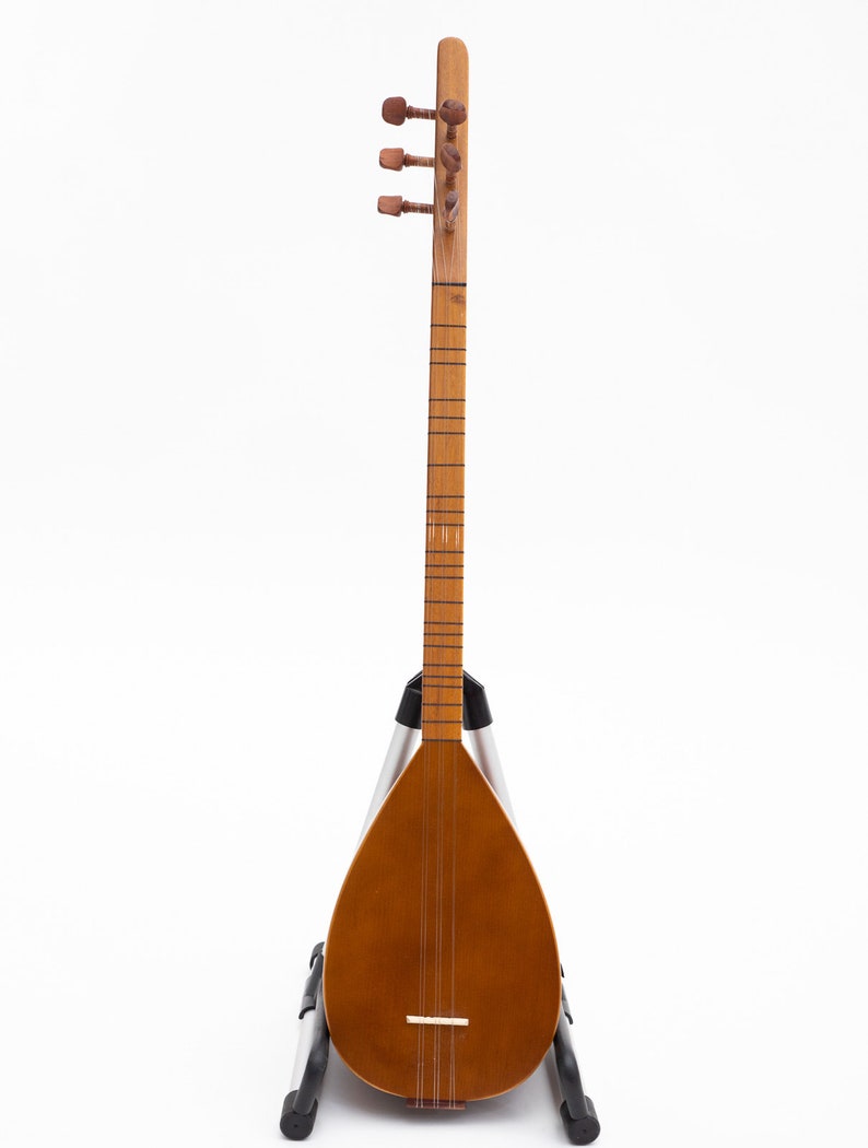 May include: A wooden stringed instrument, likely a saz or similar folk lute, with a pear-shaped body and a long neck. The instrument is a warm brown color and is supported by a silver and black stand. The headstock features tuning pegs.