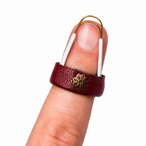 May include: A dark red textured leather ring featuring a small, round, antique brass-toned metal embellishment with a floral pattern. The ring is secured by a thin, flexible gold-toned wire loop with white plastic tips that extend upward.