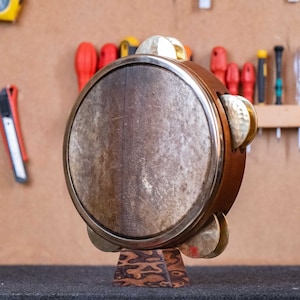 Op de afbeelding: Een houten tamboerijn met een natuurlijke huid en messing belletjes. Het instrument staat op een decoratieve houten standaard. De houten lijst van de tamboerijn is bruin en de belletjes zijn goudkleurig.