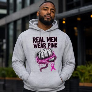 May include: A light gray hooded sweatshirt featuring the black text REAL MEN WEAR PINK above a graphic of a clenched fist wrapped in a pink chain, with a pink awareness ribbon positioned below the fist on the front of the garment.