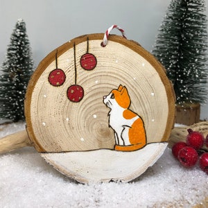 Chat roux et blanc assis dans la neige sous des boules de Noël avec des flocons de neige qui tombent.