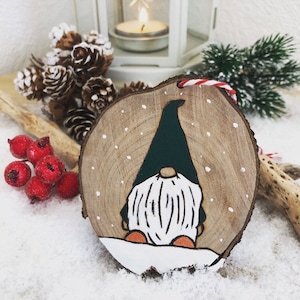 Peut inclure: Ornement de Noël en bois avec un motif de gnome peint. Le gnome a un chapeau vert foncé, une barbe blanche et des chaussures orange. L'ornement est rond avec un grain de bois naturel et une ficelle rouge et blanche pour l'accrocher. Des flocons de neige sont peints sur le bois.