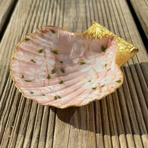 以下が含まれることがあります： 金色の縁取りと白い花柄のピンクの貝殻。貝殻は繊細な花柄で描かれており、縁に金箔のアクセントが施されています。