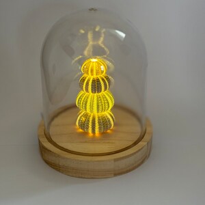 May include: A glass dome with a wooden base containing a stack of three illuminated sea urchins. The urchins are arranged in a pyramid shape and emit a warm yellow glow.