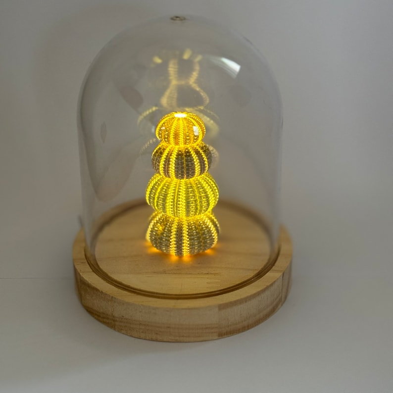 May include: A glass dome with a wooden base containing a stack of three illuminated sea urchins. The urchins are arranged in a pyramid shape and emit a warm yellow glow.
