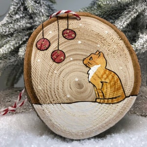 Chat tigré roux assis dans la neige sous des boules de Noël avec des flocons de neige qui tombent.