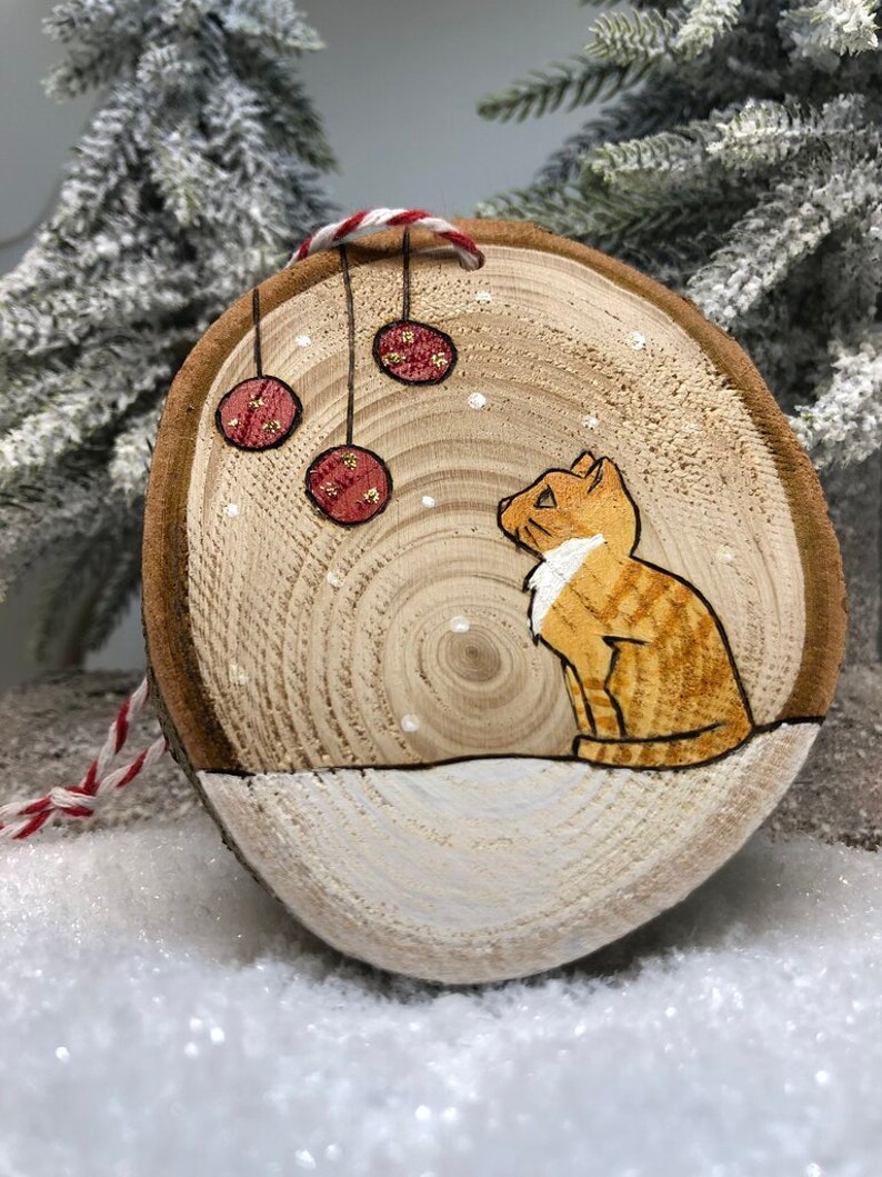 Chat tigré roux assis dans la neige sous des boules de Noël avec des flocons de neige qui tombent.
