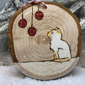 Chat blanc assis dans la neige sous des boules de Noël avec des flocons de neige qui tombent.