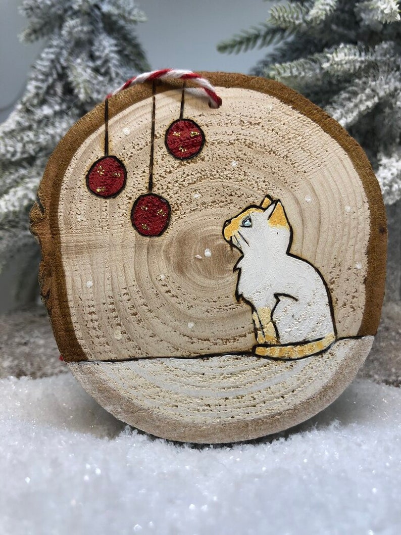 Chat blanc assis dans la neige sous des boules de Noël avec des flocons de neige qui tombent.