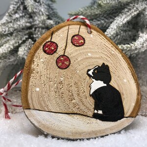 Chat noir et blanc assis dans la neige sous des boules de Noël avec des flocons de neige qui tombent.