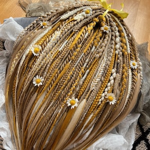 Puede incluir: Una colección de rastas sintéticas rubias, marrones y amarillas, adornadas con pequeños acentos de margaritas blancas. Las rastas están atadas con una cinta amarilla. El cabello está peinado en un patrón en capas.