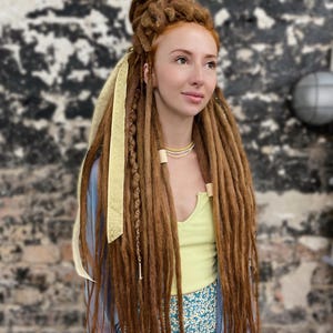 Puede incluir: Una persona con largas rastas de color marrón rojizo peinadas con un moño y mechones sueltos. El cabello está adornado con una cinta dorada y cuentas de madera. La persona lleva un top verde claro y una falda floral azul.