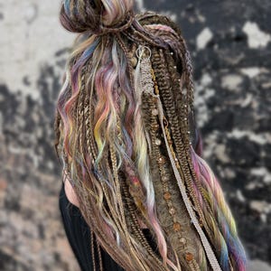 May include: A close-up of brown dreadlocks with colorful pastel highlights, styled in a bun. The dreads are adorned with decorative beads and silver charms, creating a bohemian aesthetic. The hair is styled against a blurred background.