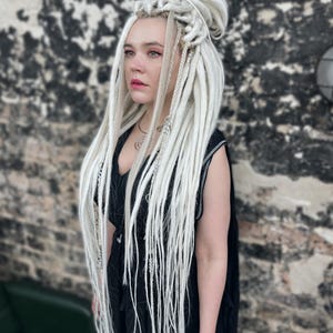 May include: A person with long, white dreadlocks styled in a high bun and loose strands. The person is wearing a black sleeveless top and standing in front of a textured brick wall. Silver jewelry is visible.