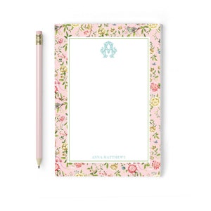 May include: A pink notepad with a floral border and a white interior. The notepad has a green border and the text "ANNA MATTHEWS" at the bottom. The notepad has a blue monogram in the center.