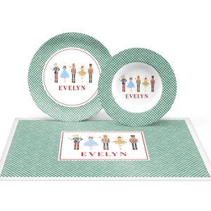 May include: A children's tableware set featuring a green and white checkered pattern. The set includes a plate, a bowl, and a placemat, all adorned with illustrations of toy soldiers and ballerinas. The name "EVELYN" is printed in red.