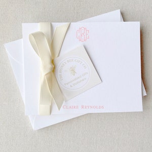 May include: A stack of white stationery cards with a pink monogram "CKE" and the text "CLAIRE REYNOLDS". A white ribbon is tied around the cards with a small white tag that says "THE HONEY BEE GIFT CO. Personalized Gifts & Stationery" with a bee illustration.