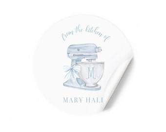Puede incluir: Una pegatina redonda blanca con una ilustración de una batidora de cocina azul y blanca. El texto "from the kitchen of" está por encima de la batidora y el texto "MARY HALLI" está por debajo de la batidora.
