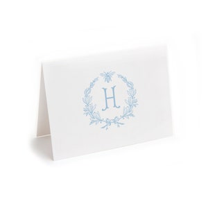 May include: A light blue card with a wreath of leaves and a bee, with the letter "H" in the center.