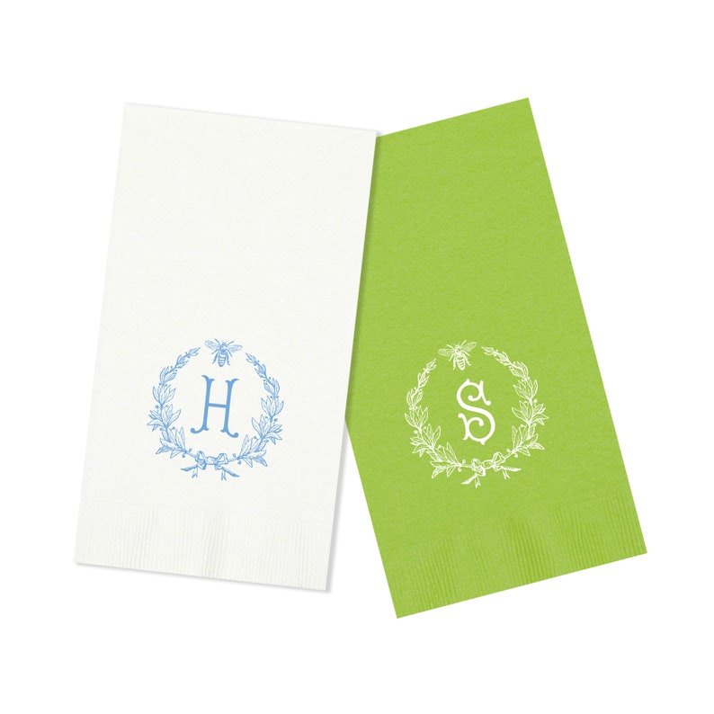 May include: Two white linen guest towels with blue and green embroidery. The towels feature a wreath with bees and a monogram. The towel on the left has the letter "H" in blue and the towel on the right has the letter "S" in white.