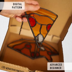 Puede incluir: Una porción de pizza de vidriera con secciones naranjas, rojas y blancas, dentro de una caja de cartón. La porción de pizza tiene un marco de metal negro y una cadena para colgar. El texto en la caja dice "DIGITAL PATTERN" y "ADVANCED BEGINNER".