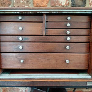 May include: A rectangular wooden machinist chest featuring a dark brown finish and multiple drawers of varying sizes. The chest includes two columns of drawers, with two on the left and four on the right, all equipped with small, round metal knobs.