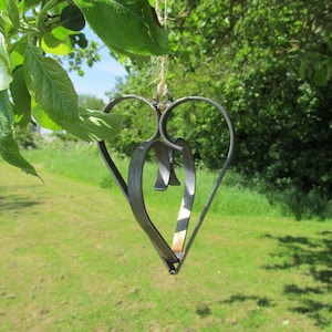 May include: A metal heart-shaped ornament hangs from a tree branch. The ornament is made of two intertwined hearts, one slightly larger than the other. The ornament is a dark gray color and has a rustic finish.