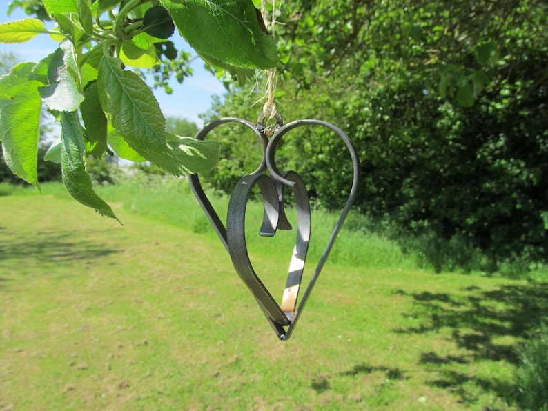 May include: A metal heart-shaped ornament hangs from a tree branch. The ornament is made of two intertwined hearts, one slightly larger than the other. The ornament is a dark gray color and has a rustic finish.