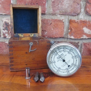 May include: A vintage wooden box with a blue velvet lining, a silver speed indicator, and metal components. The speed indicator, marked "ELOTT SPEED INDICATOR N° 2545", is displayed with the box and other parts on a wooden surface, against a brick wall.