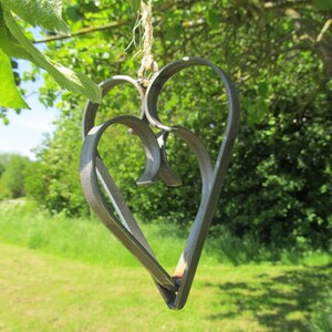 May include: A metal heart-shaped ornament with a rustic finish, hanging from a twine string. The ornament is made of two intertwined hearts, creating a layered effect. The background is a blurred image of green foliage.