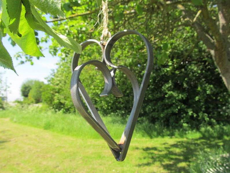 May include: A metal heart-shaped ornament with a rustic finish, hanging from a twine string. The ornament is made of two intertwined hearts, creating a layered effect. The background is a blurred image of green foliage.