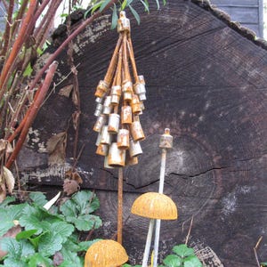 May include: A decorative metal sculpture featuring a cluster of bell-shaped ornaments with a rusted finish. Two mushroom-shaped garden ornaments with an orange-brown, textured surface are also visible. The background includes a large, dark wooden log and green foliage.