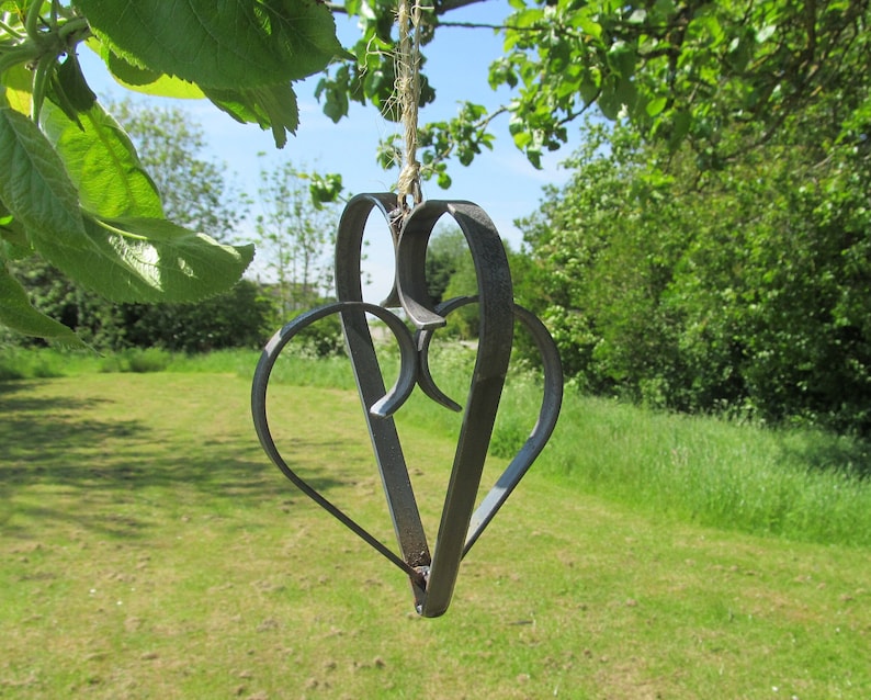 May include: A metal heart-shaped wind chime hangs from a tree branch. The chime is made of two intertwined hearts, creating a delicate and intricate design.