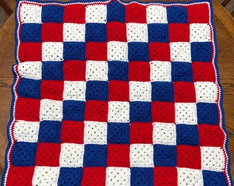 May include: A handmade crochet blanket with a checkerboard pattern in red, white, and blue. The blanket has a scalloped edge with a white border and a blue trim. The blanket is placed on a wooden table.