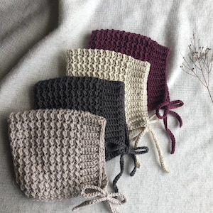 May include: Four crocheted hats in different colours: beige, brown, cream, and burgundy. The hats are made with a textured stitch and have ties at the back.