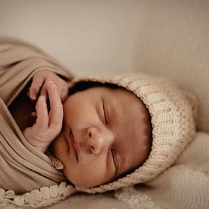 May include: A newborn baby is sleeping soundly wrapped in a beige swaddle blanket with a white crocheted trim. The baby is wearing a cream-colored crocheted hat.