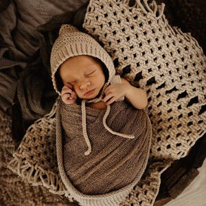 May include: A newborn baby is sleeping soundly in a brown crocheted wrap, wearing a beige crocheted bonnet. The baby is lying on a beige crocheted blanket with fringe.